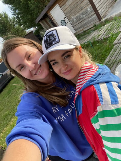 Two women are taking a selfie outdoors. The woman on the left has long brown hair and is wearing a blue sweatshirt. The woman on the right has blonde hair, is wearing a white baseball cap with a black panther logo, and a colorful striped jacket. They are smiling and appear to be in a rural or backyard setting. In the background, there is a wooden structure, some trees, and a grassy area. This scene is consistent with the exterior of a house or dacha in a dacha settlement like the one in Garevaya, Russia.