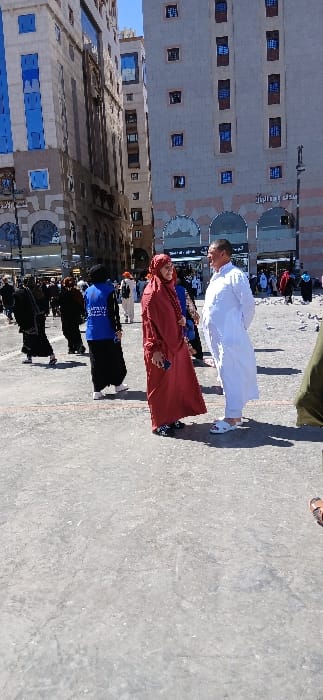 The image captures an outdoor urban plaza scene in Medina, Saudi Arabia, under bright daylight. In the foreground, a woman wearing a dark red abaya and matching hijab stands, holding a dark-colored smartphone in her left hand, facing a man dressed in a white thobe and white sandals. The man has his hands clasped behind his back. To the left of the central figures, a person in a blue vest over dark clothing is partially visible, facing away from the camera. Numerous other individuals, many dressed in traditional Islamic attire, are walking in the midground and background. The background features several tall, multi-story buildings, including one on the left with a modern facade and vertical blue-tinted windows, and another on the right with a lighter-colored facade, multiple rectangular windows, and arched ground-floor entrances bearing Arabic script. The ground is a light grey paved surface with visible cracks. A clear blue sky is overhead.