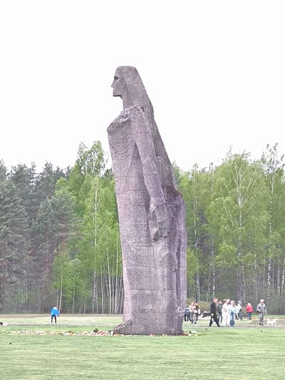 The image shows a large, gray, monolithic statue of a human figure, viewed in profile, standing in a grassy field. In the background, there is a forest consisting of both evergreen and deciduous trees. Several people are walking on the grass in small groups. The statue is located in Mežezeri, Latvia.