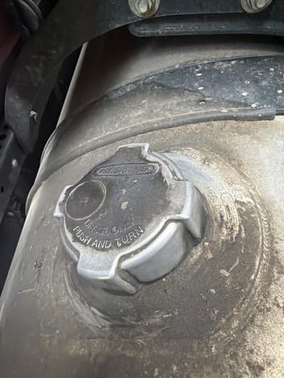 A close-up, eye-level shot captures a dusty, metal diesel fuel cap on a truck. The cap, featuring the "FREIGHTLINER" logo and instructions to "DIESEL ONLY PUSH AND TURN," is set against a grimy, cylindrical fuel tank. Faint inscriptions on the cap read, "MEETS ALL FMCSA & SID TANK MLS BY SWE," and "MFG. DA". The surrounding area is obscured by dark, shadowy components, likely part of the truck's undercarriage. The scene suggests a maintenance or operational context for a commercial vehicle.