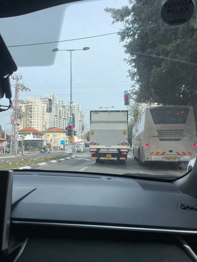 A view from inside a car shows a busy city street scene. A bus and a truck are directly ahead, stopped at a red traffic light. Buildings rise in the background, suggesting an urban area. Streetlights and power lines are visible, adding to the urban landscape. The Toyota Relax sticker is visible on the windshield. The car's dashboard fills the bottom half of the frame.