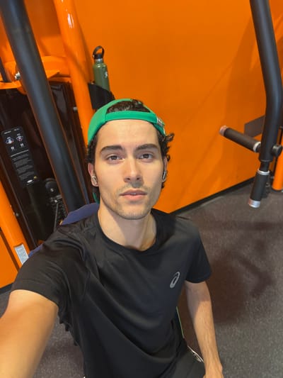 A young man with dark curly hair, wearing a black t-shirt, black shorts, a green baseball cap, and a silver earbud, stands in a gym with orange walls. He is looking at the camera and appears to be taking a selfie. Behind him, to the left, is a piece of gym equipment with instructions for use. To the right, a green water bottle is attached to the equipment. The floor is dark grey. The location is the Seraing, Belgium municipal sports complex.