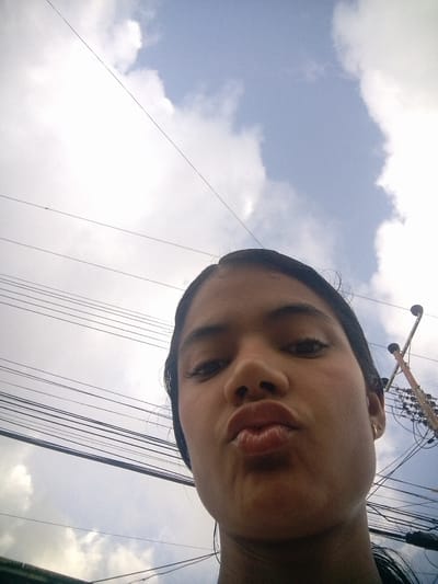 A young female individual is captured in a close-up selfie, with her face angled towards the camera, pursing her lips as if to give a kiss. She has dark hair, prominent eyebrows, and visible eyelashes. She is wearing a small gold stud earring in her visible ear. The background is a cloudy sky with visible power lines crisscrossing the frame, suggesting an urban or semi-urban environment. In the upper right portion of the image, a portion of a utility pole with some electrical equipment is visible. The overall lighting suggests it is daytime. The location is identified as El Maco, Venezuela.