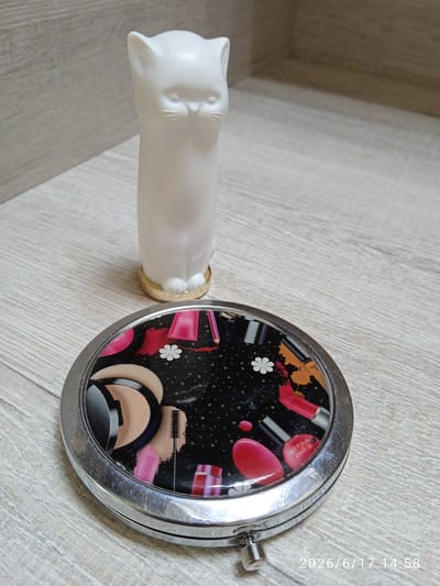 A white, stylized cat figurine stands on a light-colored wooden surface. In front of the figurine, a round, silver compact mirror is placed. The mirror's lid features a black background decorated with various cosmetic items, including lipstick, nail polish, and what appears to be foundation or powder, along with floral accents. The background is a wooden surface with visible grain. A timestamp in the bottom right corner reads "2026/6/17 14:58". There are no people or specific identifiable locations within the city of Yevpatoriya, Ukraine, visible in this image.