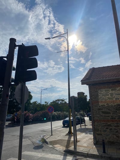 This media file captures a street scene in Thessaloniki, Greece, with a focus on a traffic light and the sky. The image is oriented vertically, with the sky dominating the upper portion. Sunlight streams through a partly cloudy sky, casting lens flare and illuminating wispy clouds.

In the foreground, a dark traffic light with three lenses is visible, with the red light illuminated. Below it, another section of a traffic light or pole is present. A metal pole extends upwards, supporting street lighting infrastructure.

The middle ground reveals a street with parked cars and a sidewalk. The sidewalk is paved with tactile paving stones, indicating it's a pedestrian area. A building with a red tiled roof and stone walls is visible on the right, with trees and foliage lining the street. A "No Parking" sign with a blue circle and red cross is displayed, along with a green traffic light further down the road. The sun's position suggests it is daytime. The overall scene conveys a sense of urban infrastructure meeting natural elements. No people are clearly visible in the image.