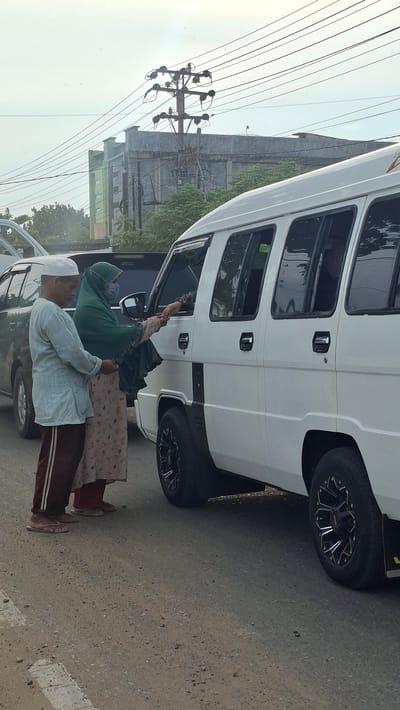 In Keudebayu, Indonesia, two individuals, an adult male wearing a white cap and a light blue long-sleeved shirt with dark striped pants, and an adult female wearing a green hijab and patterned skirt, are standing on a road next to a white van. The female is extending her hand through the open driver's side window of the van, and a hand from inside the van is reaching out to her. The male is standing to the left of the female, looking down. The road surface is asphalt with scattered debris and white markings. In the background, there are other vehicles, buildings, trees, and utility poles with numerous overhead wires. The sky is overcast.