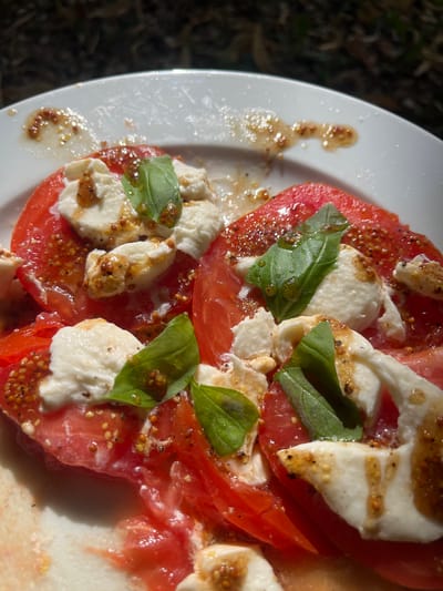 A close-up, overhead shot shows a caprese salad served on a white plate. The salad features sliced red tomatoes topped with fresh mozzarella cheese and garnished with basil leaves. A vinaigrette dressing, appearing to be olive oil-based with whole mustard seeds, is drizzled over the ingredients. The plate is placed outdoors, with dappled sunlight suggesting it is on a table in a garden or patio area. The background is out of focus, showing dark foliage. No people are visible. The location is Bièvres, France.