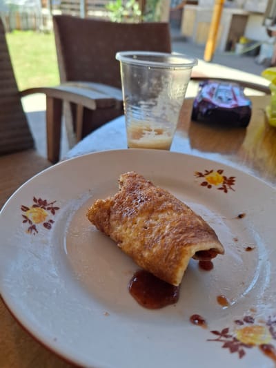 A close-up shot focuses on a single, partially eaten rolled pancake or crepe on a white plate with floral decorations. The food item has a golden-brown, slightly uneven surface and appears to be filled with a dark red jam or fruit filling that has spilled out onto the plate. Several small droplets of the same jam are scattered around the food. The plate is positioned on a wooden table. In the blurred background, a clear plastic cup with remnants of a beverage is visible, along with a toy car. Further back, a wooden chair and a grassy area suggest an outdoor or semi-outdoor setting, possibly a backyard or patio. The overall scene implies a casual meal or snack, with the toy car and outdoor elements hinting at a family-friendly environment. The image does not contain any identifiable landmarks specific to Galabovo, Bulgaria, beyond the general context of a domestic setting.