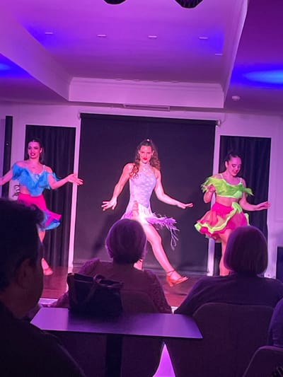 An indoor entertainment venue in Calvià, Spain, features a live dance performance. Three adult female dancers are on a stage with a dark background, illuminated by artificial purple and blue stage lighting. The dancer on the left wears a blue fringed top and a pink skirt, posing with extended arms. The central dancer wears a light purple patterned leotard with leg fringe and brown dance heels, mid-action with one leg raised. The dancer on the right wears a bright green fringed top and a neon pink and green ruffled skirt, also posing with extended arms. All dancers are smiling. In the foreground, several audience members are seated at dark tables, viewed from behind, observing the performance. A dark handbag rests on one of the tables. The ceiling is white with decorative molding and recessed lighting. A speaker unit is visible on the left side of the stage.