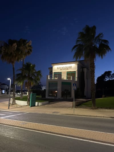 The image depicts the exterior of a modern building at night in Finestrat, Spain. The building, illuminated by internal and external lighting, features a white facade with green accents around windows and balconies. The signage on the building reads "MUERCIELAGO GRUPO." In front of the building, there is a paved walkway, a pedestrian crossing, and a road. Several palm trees are visible on the left and right sides of the building, with one prominent palm tree in the foreground to the right. The sky is a deep blue, characteristic of twilight or night. A street light is visible on the left, and a pedestrian crossing sign is placed near the building's entrance. The overall scene conveys a calm, urban night environment.