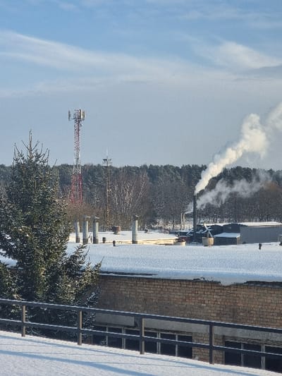 The scene depicts a daytime winter landscape in Jaunmārupe, Latvia, under a predominantly clear blue sky with wispy high-altitude clouds. The immediate foreground features snow-covered ground with a dark, multi-bar metal railing extending horizontally. To the left, a snow-dusted evergreen tree partially obstructs a single-story brick building. This building has a flat, snow-covered roof from which five cylindrical, silver-colored vents or small chimneys project vertically.
In the mid-to-background, a dense forest composed of coniferous and bare deciduous trees forms a horizon line. Above the left section of the treeline, a prominent red and white lattice cellular tower with multiple antenna platforms is visible, positioned alongside a smaller, darker communication tower. To the right, an industrial facility is present, consisting of various dark-roofed structures and a tall, dark smokestack actively emitting a thick, white plume of smoke that drifts diagonally upwards and rightward across the sky. No human subjects, animals, or active interactions are discernible within the frame.