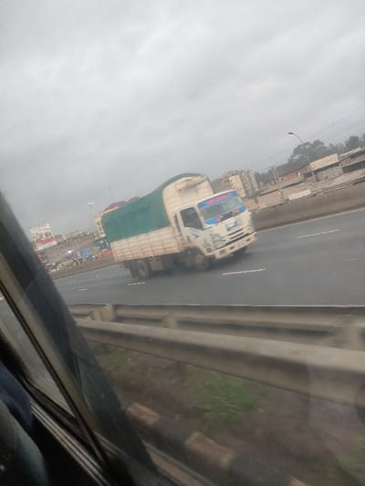 The image displays a multi-lane asphalt highway scene in Githurai, Kenya, under an overcast sky, captured from the interior of a vehicle. A white box truck, featuring a green tarp covering its cargo area and a blue and red visor on its cab, is in motion from right to left across the mid-ground. White lane markings are present on the roadway. In the immediate foreground, a dark vehicle window frame and a gray metallic guardrail are visible, with sparse green vegetation below. The background reveals urban infrastructure: on the left, a billboard with red and white graphics precedes a cluster of multi-story buildings. To the right, additional buildings, a street lamp, and distant trees are observable beyond a concrete barrier paralleling the highway. No individuals are explicitly discernible.