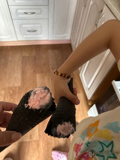 The image captures a close-up view of two black ice cream cones with pink ice cream, held by a person's hands. The person's arm, adorned with a beaded bracelet, is visible on the left. They are wearing a light-colored t-shirt with colorful star graphics, and a silver necklace with a key pendant. The scene appears to be indoors, with hints of kitchen cabinets and wooden flooring. The lighting suggests it is daytime. The context provided places the location in Urazovo, Russia. The image focuses on the ice cream cones, with the person's hands and forearms being the only visible parts of the person. The ice cream cones are dark, with a textured waffle exterior, and the pink ice cream is partially melted and exposed. The overall impression is casual, possibly a moment of enjoying a sweet treat.