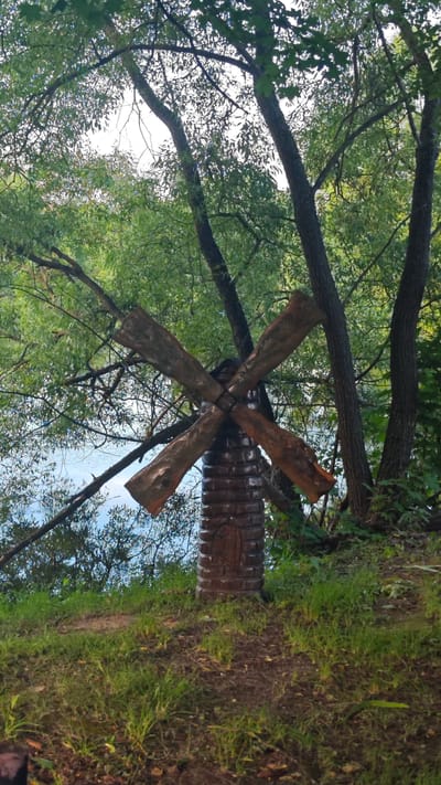 The image depicts a rustic, wooden windmill structure situated outdoors, likely in a park or natural setting near a body of water. The windmill has four large, weathered wooden blades arranged in a cross shape, attached to a cylindrical base that is carved to resemble stacked bricks. The base also features a small, recessed decorative element.
The windmill stands on a grassy embankment overlooking a calm, blue body of water, possibly a river or lake. Behind the windmill, several mature trees with lush green foliage create a dense canopy. The branches of these trees extend into the frame, partially obscuring the sky, which appears overcast, suggesting a cloudy day. The ground in the foreground is covered with grass and fallen leaves, indicating a natural, perhaps slightly unkempt, environment.
The overall impression is one of a tranquil, bucolic scene. The style of the windmill suggests it is a decorative piece rather than a functional one. The lighting is soft, consistent with diffused daylight. The setting evokes a sense of peace and connection with nature. No people are visible in the image, and there is no discernible text. The location context of Щапово, Russia, reinforces the impression of a rural or semi-rural setting.