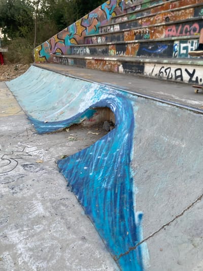 The image shows a concrete skatepark with graffiti. In the background, there are tiered concrete steps covered in colorful graffiti. The foreground features a curved concrete ramp painted with a blue wave design. The ground around the ramp is also concrete and has various graffiti markings and scuff marks. A person's legs and a skateboard are visible in the upper right corner of the image, suggesting recent activity. The environment appears to be outdoors, with some vegetation visible behind the skatepark. The overall impression is of an urban, well-used recreational space.