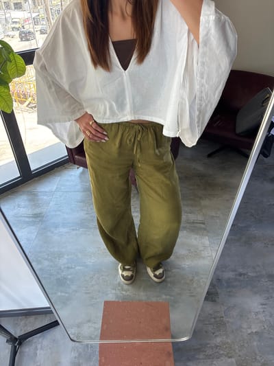 A full-body reflection of a woman in a floor-length mirror. The woman is wearing a white tunic-style top, olive green trousers, and neutral-colored sneakers. She has long brown hair and is wearing multiple rings on her left hand.

The mirror is set on a grey tiled floor, with part of a brown brick or tile visible in the corner. A burgundy armchair is visible behind the woman in the reflection. A potted plant with green leaves is visible near a window. Outside the window, a city scene is reflected, including buildings, construction equipment, and vehicles. The reflection suggests an urban environment. The location is Zovuni, Armenia.