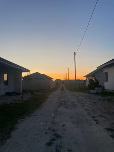 This media file captures a quiet scene at dusk in a rural area. The image depicts a dirt road winding between unfinished houses, suggesting a developing residential area. A utility pole with wires stretches across the clear, pale blue sky, which transitions to a soft orange hue near the horizon, indicating either sunrise or sunset. The overall atmosphere is peaceful and serene, perhaps even a little melancholic, with the subdued lighting and the unfinished state of the buildings. There are no people or specific activities visible, but the presence of the road and houses points to a residential setting. The location context provided is Мухавецкі сельскі Савет, Belarus. There is no visible text in the image.