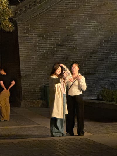 Two women are posing for a selfie in an urban alleyway at night. One woman is wearing a white top and dark pants. The other woman, slightly in front, is wearing a tan coat and blue jeans. A mobile phone is visible in the hands of the first woman. The alleyway has a brick wall on one side and a lighter wall on the other, indicating a possible corner. In the lower portion of the image, there are people sitting on the ground. The location is likely Jinzhou, China.