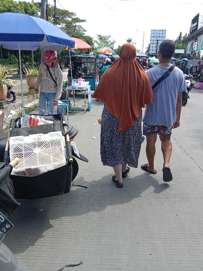 The image shows a street scene in Kendal, Indonesia (latitude: -6.933, longitude: 110.204). A man in a grey t-shirt and floral patterned shorts walks next to a woman wearing a brown hijab and a black and white floral dress. The man is looking down at the ground, while the woman is looking straight ahead. Behind them, a man in a blue shirt and a red helmet is walking away from the camera while carrying a black bag. The man with the red helmet is pushing a small cart filled with a white crate. There is a blue and white umbrella on the left side of the image, and a food stall with a yellow awning is in the background. There are several other people walking around in the background, as well as a few motorcycles. The pavement is grey, and the street is mostly empty.