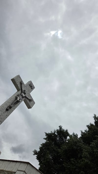 A low-angle, wide shot captures a stone crucifix with a metal sculpture of Jesus Christ, set against a cloudy sky. The crucifix is positioned in the lower-left quadrant of the frame and is oriented diagonally. A dense cluster of dark green trees occupies the lower-right portion of the image. The sky is overcast with patches of brighter light filtering through, suggesting the sun is partially obscured. The foreground shows a section of a grey, concrete structure, possibly a monument base. The scene is suggestive of a cemetery or memorial site.