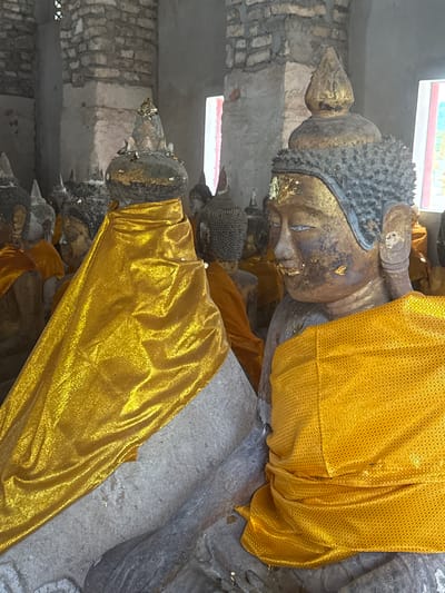 This image shows a row of Reclining Buddha statues in what appears to be a temple or sanctuary in Baan Harn, Thailand. The statues are adorned with golden robes, characteristic of Buddhist iconography. The primary subject is the central Reclining Buddha statue, with its head resting on its arm, eyes closed in a serene posture. The statue is detailed, with a textured headdress and facial features that suggest age and reverence. Gold leaf is applied to parts of the statue, including the face and headdress, adding to its sacred appearance.
Several other Reclining Buddha statues are visible behind and beside the main one, also partially covered with golden fabric. The statues are situated against a backdrop of rough stone or brick walls, and a brightly lit opening, possibly a window or doorway, is visible in the upper left. The lighting suggests an indoor setting, likely during daytime. The overall atmosphere is one of peace and spiritual devotion. No visible text is present in the image.