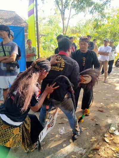 The image depicts an outdoor scene in Menjing, Indonesia, featuring a cultural gathering or ritual involving multiple individuals under bright daylight conditions.
In the foreground, a person with long, dark, wavy hair and a traditional head covering (blangkon) is positioned in a low crouch or kneel, facing away from the viewer. This individual wears a black t-shirt, a white undershirt, and a patterned gold and brown sarong. Their right hand is placed on the upper back of another male figure directly in front, while their left hand holds a white, shield-shaped object with red and brown circular patterns.
The central figure, a male, has his back towards the viewer. He wears a black long-sleeved shirt with a red collar, blue jeans, and dark sneakers. The back of his shirt displays yellow text, "COBO AMBIREJO" and "ANDIREJO". A long, dark, tail-like prop, possibly representing a horse's tail, extends from his lower back towards the ground, with a decorative base. He appears to be leaning forward, engaged with another male figure standing opposite him.
This third central figure, an older male, faces the camera. He wears a black long-sleeved shirt and black pants, topped with a traditional head covering similar to a blangkon. His right hand rests on the left shoulder of the individual with his back to the camera. Near his feet, he holds a decorated, whip-like object with yellow and green adornments.
To the left of the scene, partially visible, is a male in a grey t-shirt and white shorts, carrying a small satchel. Behind him, an older female in a light green collared shirt stands with her arms crossed, observing. Further in the mid-ground and background, several other individuals are present, some observing the central interaction, including one male in a white t-shirt and light pants with arms crossed.
The environment consists of a dry, uneven dirt and concrete ground scattered with fallen leaves and a clear plastic cup. In the background, green foliage and trees are visible, suggesting a rural or semi-rural setting. Structural elements include a partial view of a building with a tiled roof, a blue tarp or tent structure, and a tall vertical pole with a yellow and purple banner. The scene is illuminated by strong sunlight, casting distinct shadows.