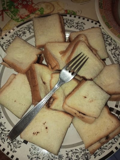 The scene depicts a tabletop in Kodarapalli, India. A round white plate, featuring a black and white floral and scrollwork border, is centrally positioned. The plate contains an elevated mound of approximately 15-20 small, uniformly cut pieces of white bread, presented as sandwiches in triangular and square forms. A dark brown filling is visible along the cut edges of the bread pieces and as minor flecks on their surfaces. A silver-colored metal fork rests diagonally across the pile of bread, with its tines pointing towards the upper right. The fork's handle is embossed with the text "STAINLESS STEEL INDIA". Portions of a multi-colored placemat with red and green patterns are visible to the left and upper-left of the plate, and a section of another dish or surface with an orange border and geometric pattern is visible to the upper right. No individuals are present in the image.