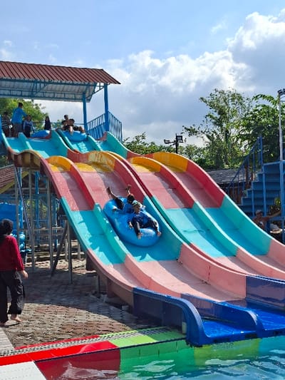 The image shows a water park slide located at a water park in Kecamatan Tulung, Indonesia. The slide is made of blue and pink plastic, with a blue and green tiled area below it. It is a double slide with two people riding on it. The people are both wearing blue shirts and blue shorts. They are riding in a blue tube and are about halfway down the slide. Other people can be seen in the background, including a woman in a black hijab standing near the bottom of the slide and a group of people on the top platform. The sky is blue and there are clouds in the sky. There is vegetation in the background to the right of the slide.