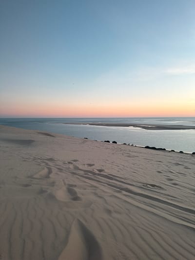 The image shows a vast sand dune in the city of La Teste-de-Buch, France, overlooking a body of water. The dune is covered in fine sand with visible footprints, indicating recent human activity. The texture of the sand shows ripples and undulations, shaped by wind. Beyond the dune, a calm body of water, likely the Arcachon Bay, stretches towards the horizon. The sky exhibits a gradient of colors, transitioning from a pale blue at the top to soft hues of pink and orange near the horizon, suggesting a sunset or sunrise. A few birds are visible as small dark specks in the sky. The environment is natural and serene, characterized by the interplay of sand, water, and sky.