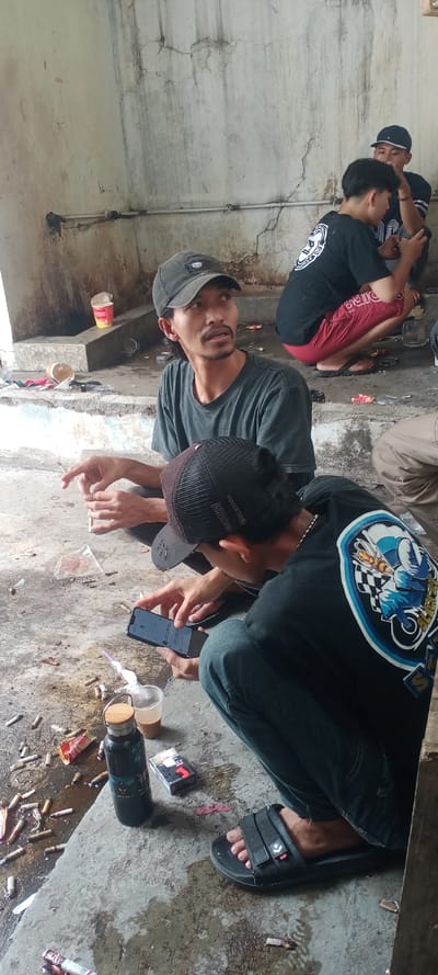 The image depicts five adult males gathered in a dilapidated, concrete-walled area in Sulaeman, Indonesia. The environment features a stained, wet concrete floor littered with numerous cigarette butts, discarded packaging, and two empty plastic cups, one containing a light brown liquid (likely coffee). A dark green water bottle with a wooden cap and a pack of Marlboro cigarettes are also on the ground.

In the immediate foreground, a male wearing a black mesh baseball cap, a black t-shirt with a blue and white graphic on the back, dark jeans, and black open-toed sandals is squatting. He is actively engaged with a black smartphone, held in both hands, with its screen visible.

To his right, another male, wearing a dark grey t-shirt and a dark baseball cap, is also squatting. He holds a lit cigarette in his left hand and looks upwards and to the right, away from the camera.

In the background, three additional males are present. One is squatting on a slightly elevated concrete platform, wearing a black t-shirt with a white graphic, red shorts, and black sandals. Another, partially obscured, wears a dark baseball cap and appears to be interacting with the man in red shorts. A fifth male is visible further back, also wearing a dark cap.

The concrete walls are marked with cracks, water stains, and discoloration. A section of white PVC piping runs horizontally across the background wall. An empty red cup noodles container sits on a low concrete ledge. The scene is illuminated by natural daylight.