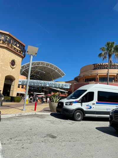 A white Ford Transit van, marked with "TA" and a wheelchair symbol, is parked near a building with "Texas Churrascaria" visible. The sky is bright blue. The entrance to the Dolphin Mall is apparent, marked with its name in large letters. People are visible under the mall's awning. A lamp post and a circular structure are notable features.