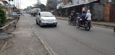 A silver sedan drives down a paved road in Keshero, Democratic Republic of the Congo. The road is in fair condition, with some cracks and potholes. The sidewalk to the left of the road is uneven and dirt, while the sidewalk on the right is paved and in good condition. A small truck is parked in the background on the left side of the road, and several people are visible, some on foot and others driving motorcycles. The scene is typical of a city in the developing world, with a mix of old and new vehicles and some signs of neglect in the infrastructure. There is a brick building and a few trees in the background on the right side of the image. The driver of the silver sedan is focused on the road. Three people are riding a red and black motorcycle toward the left of the image, the passenger on the back is wearing a white hooded sweatshirt.