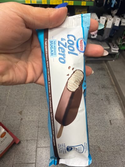 A person's hand holds a Nestlé Cool & Zero ice cream bar. The ice cream bar is light blue and white with an image of the ice cream on the front. The hand has manicured nails with blue polish. The background is blurred and appears to be inside a store. The location is Ravno pole, Bulgaria.