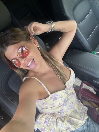 A woman is sitting in the driver's seat of a car, looking at the camera and sticking out her tongue. She is wearing sunglasses, a floral top, and a necklace. Her hair is blonde and pulled back. The car interior is visible, with black leather seats. A burgundy handbag is on the passenger seat. There is no information in the image to suggest a location in the city of Пастухово, Russia.