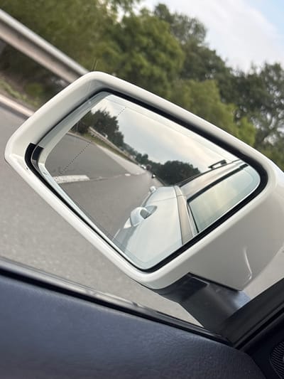 This media file captures a view from inside a vehicle, focusing on the exterior side mirror. The mirror reflects a road with a white line marking, suggesting the vehicle is in motion on a paved surface, likely a highway or main road. Trees and foliage line the side of the road, indicating a rural or semi-rural setting. The sky appears overcast or hazy, casting a soft light over the scene. The vehicle's white exterior is visible around the mirror housing. The interior of the car, specifically a dark, textured surface, is visible in the foreground and right side of the frame, implying the photograph is taken from the driver or passenger side. The overall impression is that of a journey taking place during daylight hours, with no specific time of day or weather conditions beyond the soft lighting. The location context is Gorna Vasilitsa, Bulgaria.
