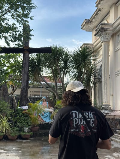 The image shows a person wearing a black t-shirt with a "Dark Place Tattoo and Barbershop" logo, and a beige baseball cap, facing away from the camera. They are standing in a courtyard with wet paving stones, suggesting recent rain. To the left, there is a large wooden cross and a statue of Mary holding a reclining figure, likely Jesus. In the background, there are palm trees and a building with classical architectural elements, possibly a church or a colonial-era structure. A sign with Vietnamese text is visible near the statue. The sky is partly cloudy and a light blue. The location is identified as Diên Điền, Vietnam.