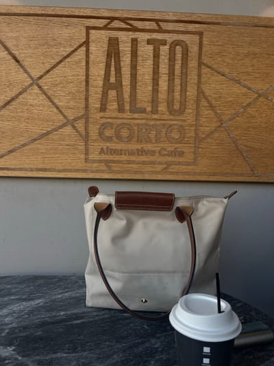 An indoor scene at Alto Corto Alternative Cafe, located at G. Papadopoulou 1, Agia Paraskevi, Greece. A rectangular wooden sign with the engraved text "ALTO CORTO Alternative Cafe" is mounted on a light grey wall, occupying the upper portion of the frame. Below the sign and wall, a dark grey marble-patterned tabletop is visible. On this tabletop, a light beige nylon tote bag with brown leather trim and handles rests to the left. To its right, a black disposable coffee cup with a white lid and a black straw is positioned. A metallic object is partially visible behind the coffee cup to the right. No people are depicted.