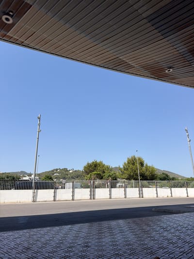 The image shows a clear blue sky above a street and a building. On the left side of the image, a portion of a building with corrugated metal siding is visible. A round object, possibly a light fixture or camera, is attached to the siding. In the center and right of the image, a paved road runs alongside a sidewalk with a decorative tiled pattern. A metal fence with concrete supports separates the road from a vegetated hillside. Trees with dense green foliage cover the hillside, and white buildings are scattered amongst them. Several streetlights with multiple fixtures are positioned along the road. The scene appears to be set in Sant Antoni de Portmany, Spain, under clear, sunny weather conditions during the daytime. There is no visible text in the image.