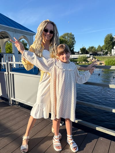 An adult female and a young female stand on a dark brown wooden deck, posing for a photograph under clear blue skies in Dumino, Russia. The adult female, positioned slightly behind and to the left of the younger female, has blonde dreadlock-style hair, wears dark oval sunglasses, a yellow and white checkered shirt over a white top, a white tiered dress, and white sandals. She forms a peace sign with her left hand and has her right arm extended behind the younger female. The young female, with brown hair and bangs, wears a long-sleeved white dress with a dark dotted pattern and white sandals. She forms a peace sign with her left hand and holds a silver metal railing with her right hand. Both individuals are smiling directly at the camera.
The metal railing separates the deck from a body of water, where several ducks are visible in the midground. In the left background, a white structure with a blue gabled roof and arched openings is partially visible. The background also includes green trees, shrubs, and a landscaped area with large rocks across the water. Further in the distance, white buildings, including one with a visible dome, are present. The scene is brightly illuminated by sunlight, casting distinct shadows on the wooden deck.