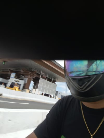 A first-person perspective shot captures a person wearing a black t-shirt and a black motorcycle helmet with a iridescent visor. The person is on a roadway, likely passing through a toll plaza, with a blurred view of the toll booths and surrounding infrastructure. The background features the architecture of a modern building or structure, possibly a commercial establishment or part of the toll plaza complex, with clear skies visible. The image appears to be taken on a sunny day in South Denpasar, Indonesia, given the visual cues of a developed urban environment and typical Indonesian architecture.