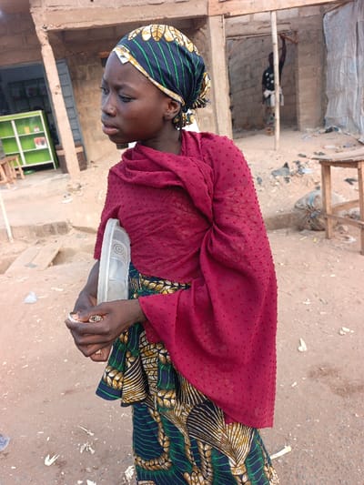 A young woman stands outdoors, dressed in a patterned headscarf, a dark red shawl, and a colorful skirt. She holds a clear plastic container in one hand and something small in the other. Behind her, there are buildings, including one with a green cabinet visible inside. A person stands in the background near a wooden structure. The ground appears dusty, with scattered debris.