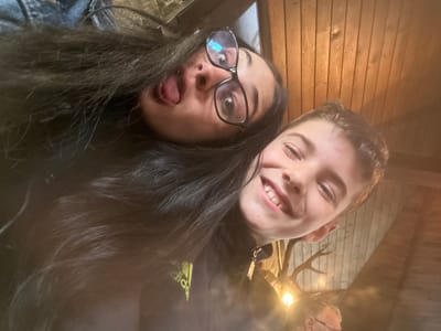 Two people are visible in a close-up selfie-style shot. One person has short light-colored hair and is smiling. The other person has long dark hair, glasses, and is sticking their tongue out. The background features wooden paneling and a decorative antler fixture with a lightbulb. Part of a garment with an Adidas logo is visible. The location is Sadievo, Bulgaria.