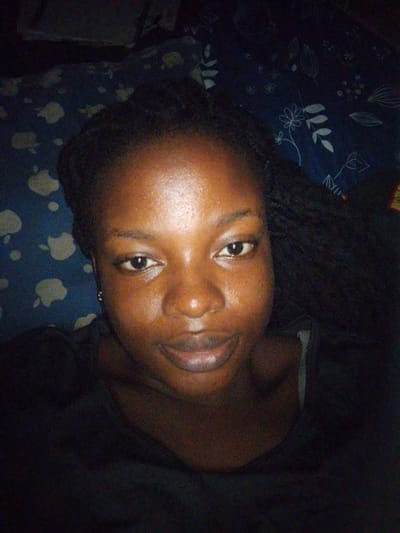 A close-up, overhead shot shows a person with dark skin and braided hair. The person is wearing a dark shirt and appears to be lying down on a patterned surface with light-colored abstract shapes and floral designs in shades of blue and white. The lighting is dim, casting shadows across the person's face, which has a slight sheen, possibly from sweat or product. The person's eyes are open, and their mouth is closed, with a hint of a smile. The background appears to be a bed or a similar soft surface. The image has a grainy texture, typical of low-light photography. There is no clear indication of a specific location within the city of Evbogida, Nigeria, other than the general suggestion of an indoor setting.