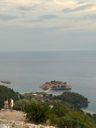 This vertical, medium-shot photograph captures a scenic view of the Adriatic Sea and a coastal village in Montenegro. Two individuals, a man and a woman, stand on a rocky cliff overlooking the water. The man is wearing a light-colored short-sleeved shirt and khaki pants, and the woman is in a light-colored dress. They are positioned towards the right side of the frame, on a rocky outcrop.

In the distance, a picturesque island settlement with red-tiled roofs is visible, connected to the mainland by a narrow causeway. The sea is a calm, blue expanse, with a few small boats dotting its surface. Lush green trees and vegetation cover the surrounding hillsides and the island itself. The sky is a mix of soft clouds and a hazy blue, suggesting it might be late afternoon or early morning, with diffused sunlight. The overall atmosphere is peaceful and serene, with a sense of awe inspired by the natural beauty and the charming architecture. There is no visible text in the image.