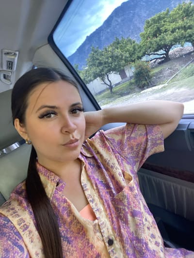 A woman is seen from the chest up, sitting inside a vehicle. She is wearing a light-colored, patterned button-up shirt with purple and orange accents, and a peach-colored tank top underneath. Her left arm is bent, resting on her raised left elbow, with her hand behind her head. The woman has long dark hair pulled back into a ponytail, prominent false eyelashes, and is looking towards the camera with her lips slightly pursed. The background, visible through the vehicle's window, shows a mountainous landscape with green trees and a clear blue sky. The interior of the vehicle is also visible, including a headrest and a part of the dashboard. The image was taken in the city of Irganai, Russia.