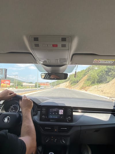 The image shows the interior of a car, viewed from the perspective of the driver. The driver's hands are on the steering wheel, and their arms are visible. The dashboard of the car is also visible, including the infotainment system, which displays song information. Outside the car, a road can be seen stretching ahead, with construction work in progress on the sides. There are trees and hills in the background. The sky is blue and partly cloudy. A sign advertising "Drive In & Out" is visible on the left side of the road. The location is identified as Lastva Grbaljska, Montenegro.