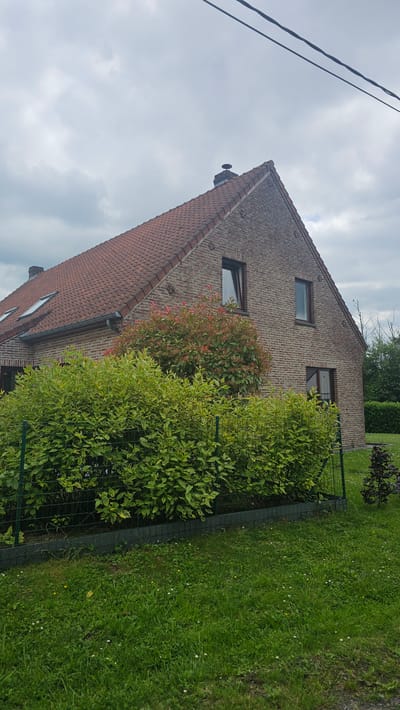 This is a medium-shot, eye-level, outdoor, daylight, full-color photograph of a brick house with a red tile roof. The house is located in Boussu-lez-Walcourt, Belgium. A large green hedge runs across the front of the property, behind a green wire fence. The sky is overcast with some clouds. There are power lines visible in the upper portion of the image.