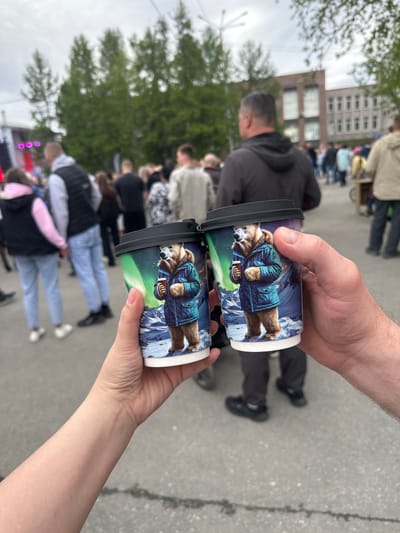 Two paper coffee cups are held up by two different people in a crowded outdoor setting. The cups feature a picture of a brown bear wearing a blue parka and holding a red book or object, against a background of snow-capped mountains and the Aurora Borealis. The person holding the top cup appears to have a darker skin tone than the person holding the bottom cup. The background shows a cityscape with a brick building, trees, and multiple people milling around. Based on the information in the image title, this event is taking place in the city of Apatity, Russia. The presence of many people suggests a public event or gathering.