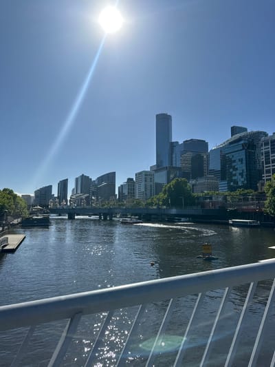 A cityscape stretches under a clear blue sky, punctuated by a bright sun. Tall buildings line a river, reflecting sunlight and suggesting a bustling urban environment. Part of a bridge railing is visible on the right, hinting at the viewpoint. The river shows signs of activity, with possibly a boat's wake or some other disturbance.