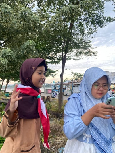 The scene depicts an outdoor setting in Kalitengah, Indonesia. Two females are positioned in the foreground. On the left, a young female, estimated to be a child, wears a dark brown hijab and a brown uniform adorned with a red and white sash. She is smiling, looking towards her right, and has her left hand raised with two fingers extended in a "V" gesture. On the right, an adult female wears a light blue patterned hijab, a light blue long-sleeved top with white bows, and a white lower garment. She is wearing light-rimmed glasses and is focused on a light green smartphone held horizontally with both hands.

The background features lush green and variegated foliage of trees, with visible tree trunks and branches. Ground cover consists of sparse grass, dirt, and several small log stumps. To the left, a body of water is partially visible. Further back, a series of simple structures, possibly market stalls or small shops, are visible along what appears to be a pathway or street. Two motorbikes are parked in the distance near these structures. Overhead, power lines traverse a pale, cloudy sky.